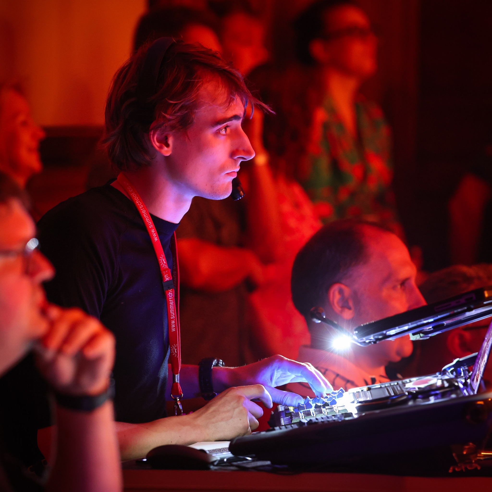 The image shows Josh working diligently at a Chamsys MQ70 lighting console, which is designed for live event production. He is deeply focused on the equipment, adjusting controls and ensuring the lighting setup is perfect for the event. The console is illuminated, highlighting its various buttons and screens, which Josh is skillfully navigating.
Josh is wearing a headset with a microphone, indicating he is communicating with other team members. A lanyard with a name badge hangs around his neck, marking him as part of the event staff. The background is dimly lit with other people present, some of whom are attentively watching the event. The scene is warm, with red and orange lighting creating a vibrant atmosphere. Josh's role is crucial, as he manages the lighting effects to enhance the event's overall experience.